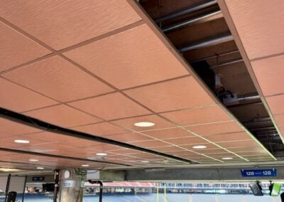 Removed Ceiling Panels Exposing Pipes and Ducts in an Indoor Stadium Concourse with Empty Bleachers Visible Beyond the Opening