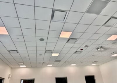 Drop ceiling Classroom with Square Tiles Recessed Lights Vents and a Ceiling mounted Projector White Walls and Three Dark Windows Along the Far Wall