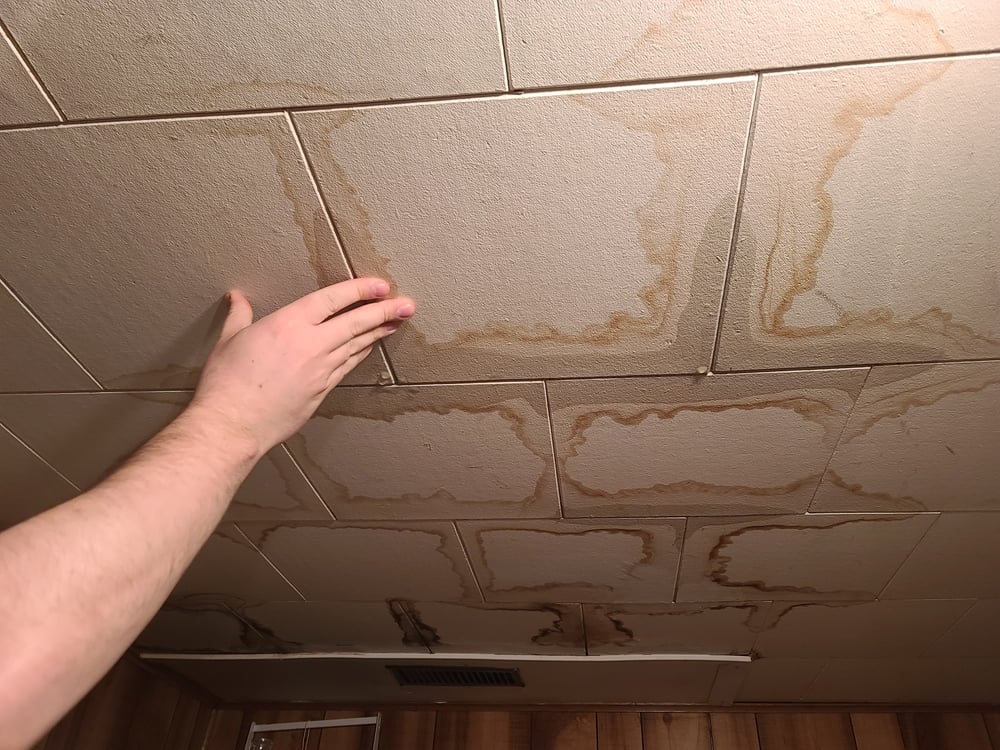 Hand pressing on a water-damaged commercial ceiling tile showing signs of moisture intrusion and deterioration.